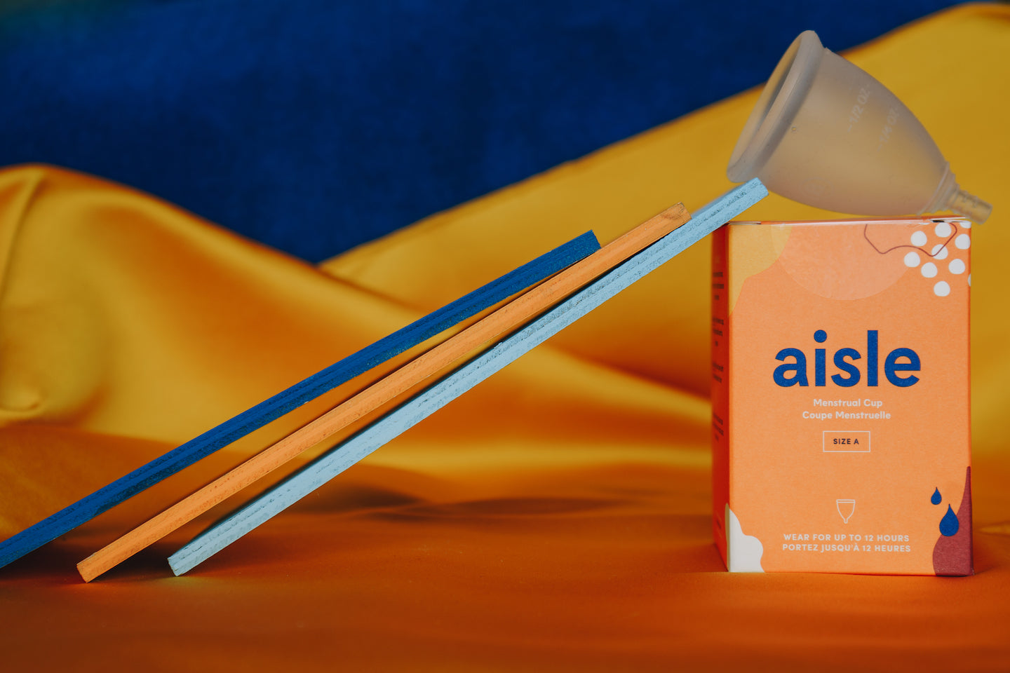 An Aisle menstrual cup sits on top of its box. There are 3 thin rectangular pieces of wood leaning up against the cup box.