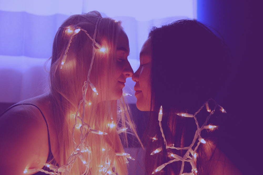 A lesbian couple touching nose-to-nose and smiling. The room is dark and they have fairy lights on their heads and shoulders.